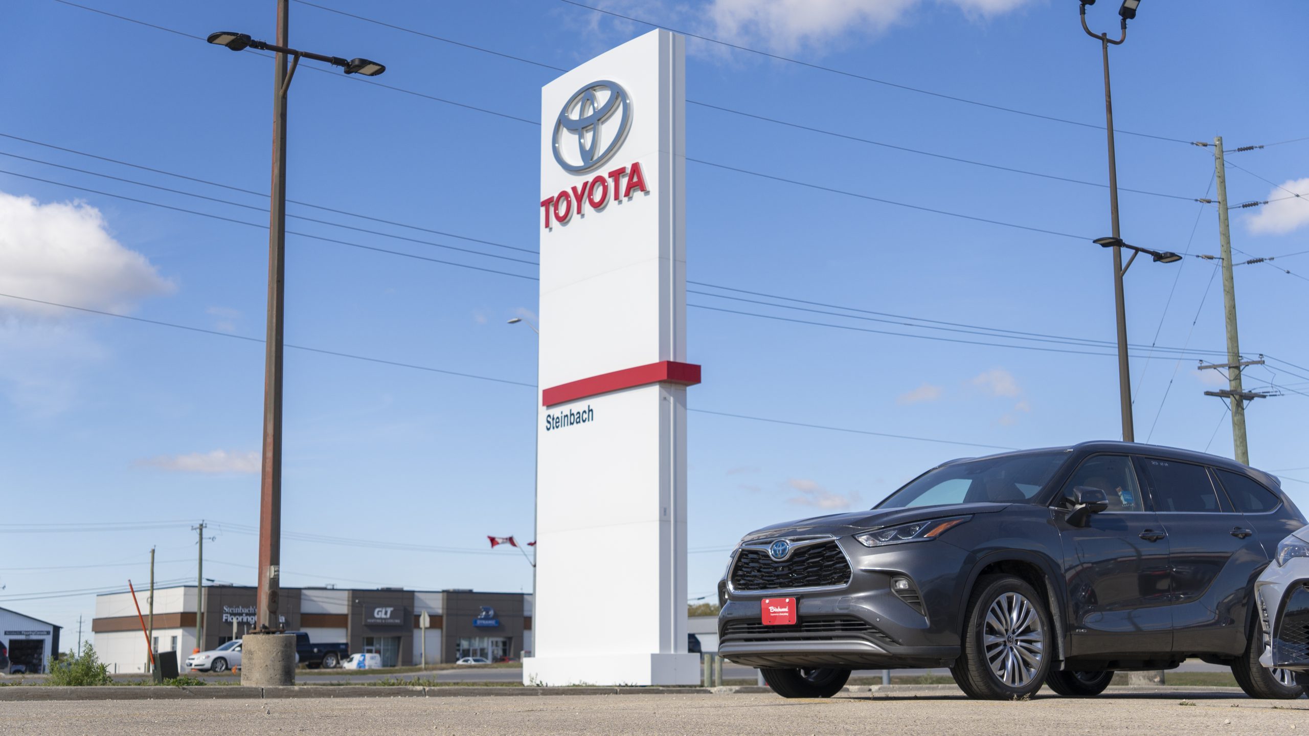 Toyota Steinbach dealership pylon sign with a grey Toyota Highlander parked in front, under a bright blue sky with light clouds.